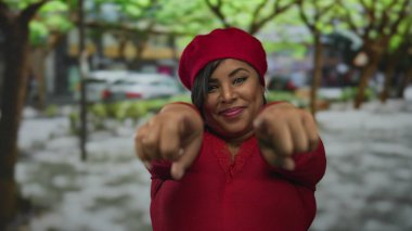 Kırmızı bereli bir kadın ağaçlarla çevrili yeşil bir parkta mutlu bir şekilde gülümsüyor ve kamerayı işaret ediyor..