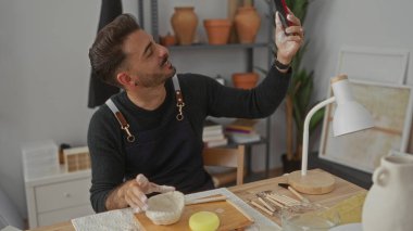 Zanaatkar atölyesinde oturan adam kırmızı akıllı telefonu tutuyor ve kil kaseyi ahşap masa üzerinde şekillendiriyor; yaratıcılık sevinci.