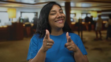 Kadın otel lobisindeki kameraya el uzatıyor. Ilık ışıklandırma ve resepsiyon masası görünür. Kendine güven..