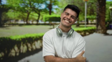 Kulaklıklı genç adam, yeşil ağaçlar ve patikalarla çevrili güneşli bir parkta gülümsüyor, rahat bir açık hava havasını yansıtıyor ve gündelik bir stili yansıtıyor..