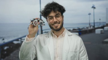 Laboratuar önlüğü giyen sakallı genç adam deniz kenarında bulutlu gökyüzünün altında, modern bilim ve teknolojiyi simgeleyen optik bir cihaz tutuyor..