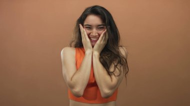 Stüdyoda turuncu bluz giyen, ağzı açık, elleri yanaklarında, kahverengi arka planı olan bir kadın. Sürpriz..