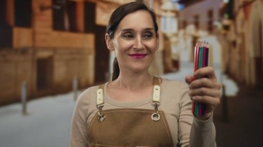 Eski kasaba caddesinde elinde renkli kalemlerle gülümseyen bir kadın. Önlük takıyor, sanat eserlerini dışarıda sergiliyor. Büyüleyici tarihi bir arkaplana karşı..