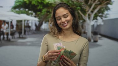 Kadın dışarıda Arjantin pesolarını sayıyor, ağaçların altında gülümsüyor, canlı bir şehir ortamında finansal başarı sergiliyor..