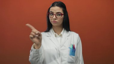 Kırmızı gözlüklü, beyaz üniformalı bir kadın kırmızı duvarın önünde işaret ediyor. Onaylamadığını belirtiyor ve bilimsel ortama odaklanıyor.