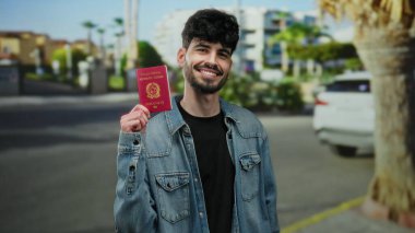 İtalyan pasaportu taşıyan kendine güvenen adam, mavi gökyüzünün altında palmiye ağaçları ve binalar olan şehir sokağında gülümsüyor..