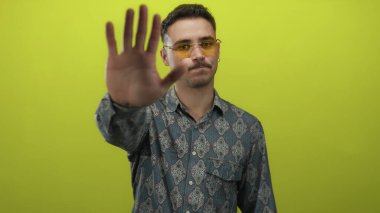 Güneş gözlüklü genç adam, parlak sarı arkaplan karşısında el işaretini uzatarak, izole bir ortamda uyarı veya reddetme mesajı iletiyor..