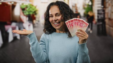 Restoran terasında gülümseyen bir kadın, elinde İsrail şekeri, parmakla gösterişli süveter giyerek para gösteriyor..