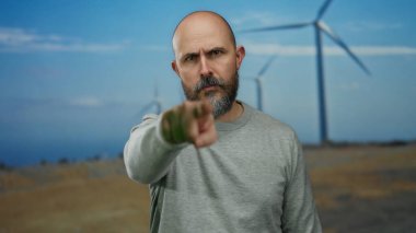 Sakallı ve kel kafalı bir adam tarlada duruyor. Arka planda rüzgar türbinleriyle gündüz vakti bulutlu gökyüzünün altında.