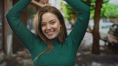 Elleriyle gülümseyen genç bir kadın şehir caddesinde yeşil süveter içinde kalp işareti oluşturuyor..