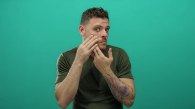 Yüzüne dokunan dövmeleri olan genç bir adam yeşil bir arkaplana dokunuyor. İlgili ifadeler takınıyor. Kişisel bakım ve cilt farkındalığı gösteriyor.