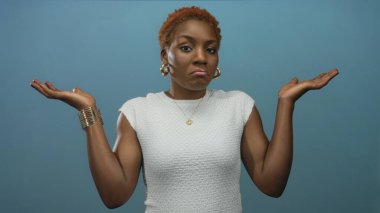 Stüdyoda omuzlarını silken ve her iki avucunu da kaldıran bir kadın, katı mavi bir duvara karşı duruyor; belirsizlik..