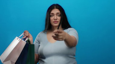 Elinde alışveriş çantalarıyla mavi duvara karşı el kol hareketi yapan genç bir kadın. Günlük kıyafetler giymiş. İletişim ve alışveriş kavramlarını ifade eden birini davet ediyor.