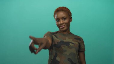 Kamuflaj üniformalı kadın stüdyodaki kamerayı işaret ediyor; işe alım motivasyonu güven güçlendirme.