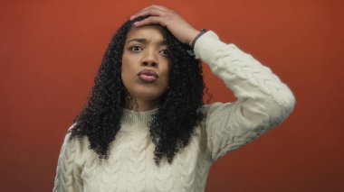 Kafa kafaya ve büzüşmüş dudaklarla turuncu stüdyo duvarına bakan kazaklı bir kadın..