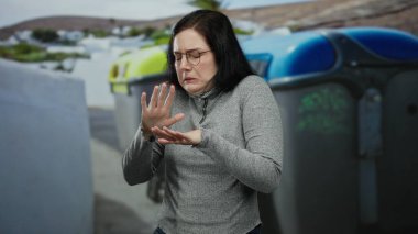 Sokak çöp konteynırlarının yanında duran esmer kadın gözlük ve gri süveter giyip şehir merkezinde rahatsız görünüyor. Açık havada duygularını ifade ediyor.