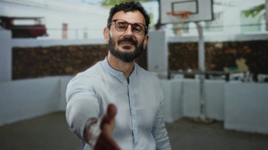 Sakallı İspanyol bir adam, samimi bir hoş geldin jesti olarak açık hava basketbol sahasında gözlüklü ve gömlekli el sıkışma teklif ediyor..