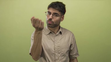 Gözlüklü ve bej renkli kısa kollu genç İspanyol adam yeşil stüdyoda avuç içini gösteriyor..