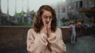 Gözlüklü ve kıvırcık saçlı genç bir kadın kalabalık bir caddede eski Roma harabelerinin önünde rahat bir süveter giyer..