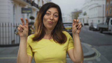 Sarı tişört giyen kadın, park halindeki araba ve binalarla dolu caddede elleri havada tırnak işareti yapıyor; şüphecilik.