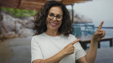 Orta yaşlı İspanyol kadın gülümsüyor ve sahile bakıyor kayalık deniz manzaralı, dışarıda neşe ve rahatlama taşıyor..