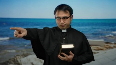 Rahip kıyafeti giyen bir adam elinde kitapla sahil kenarında duruyor ve gün boyunca açık mavi gökyüzünün altında okyanusu işaret ediyor..