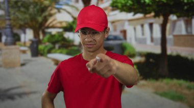 Gözlüklü, kırmızı gömlekli ve şapkalı bir adam yeşil park ağaçlarının arasında bir sokakta kamerayı işaret ediyor..