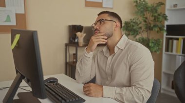 Ofis masasındaki bilgisayar monitörü ve klavye ile çenenin üzerinde dinlenen adam raflar ve saksı bitkisi arasında; yansıma.
