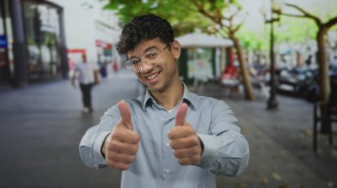 Gözlüklü ve mavi tişörtlü bir adam şehir sokaklarında enerjik bir şekilde onay veriyor..