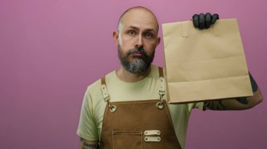 Önlüklü kel sakallı adam pembe arka planda ifadesiz yüzü olan kağıt torba tutuyor..