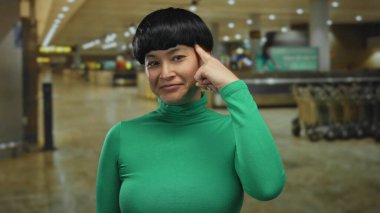 Yeşil tişörtlü genç Çinli bir kadın yoğun bir havaalanında düşünceli bir şekilde el kol hareketi yapar. Zeka ve tefekkür hissini aktarır..
