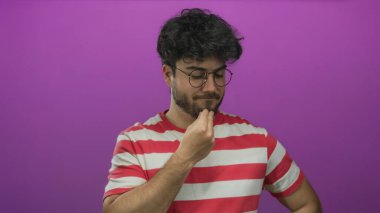 Sakallı ve gözlüklü genç adam pembe arka planda duran çizgili gömlekli düşünceli bir şekilde tefekkür ve içgözlem yapıyor..