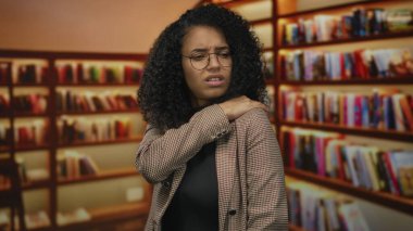 Kütüphane ortamında boynu ağrıyan bir kadın, kederli bir ifadeyle, kapalı mekanlarda kitaplıklarla çevrili, rahatsızlığı ve rahatlama ihtiyacını vurguluyor..