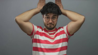 Sakallı ve gözlüklü İspanyol genç adam beyaz arka plana karşı şakacı kulaklar yapıyor, kırmızı çizgili gömlek giyiyor, neşe ve mizah taşıyor..
