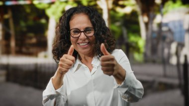 Kıvırcık saçlı ve gözlüklü olgun bir kadın şehir sokaklarında güven ve mutluluk saçan bir jest yapıyor..