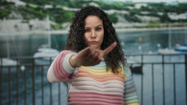 Renkli süveter giyen kadın, deniz kıyısında el hareketleriyle arka planda tekneleri olan, açık hava ortamında reddeden ya da anlaşmazlık yaşayan kadınları reddediyor..