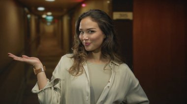 Gülümseyen kadın, aydınlık modern otel binasında kol saatini göstererek el ele uzanır; neşe içinde karşılanır.