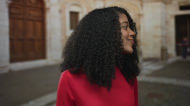 Kırmızı tişörtlü kadın eski kasabanın parke taşlı sokağında kıvırcık saçlarıyla gülümseyen yumuşak güneş ışığıyla yıkanan; mutlu keşif.