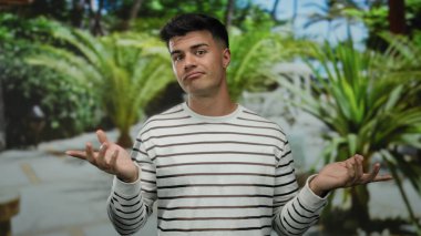 Genç adam parmaklarıyla bir açık hava parkı ortamında, yemyeşil ile çevrili, çizgili bir kazak giyiyor, oyuncu ve kendinden emin bir tavır sergiliyor..