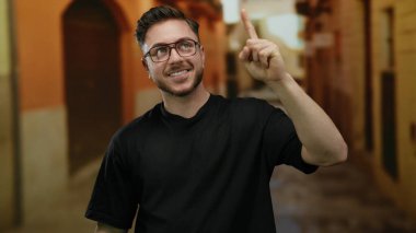 Sakallı genç adam canlı bir şehir caddesinde neşeyle yukarı bakıyor, kendine güveni tam ve şehir arkaplanının ortasında günlük kıyafetlerle çekici..