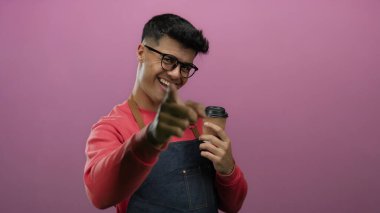 Kırmızı süveter ve önlük giymiş, pembe arka planda kahve tutan ve bardaklarla gülümseyen bir adam neşeli ve davetkar bir jest yapıyor..