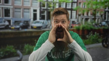 Sakallı, yeşil gönüllü tişörtlü ve elleri ağızlı bir adam sokakta sesleniyor; sosyal yardım şevki.