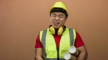 Kask ve güvenlik yeleği giymiş genç bir adam, elinde kahverengi bir arka plan, parlak kalpli profesyonelliği somutlaştıran planlar tutarken şakacı bir dille dilini uzatıyor..