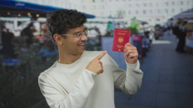 Kahvehanenin dışındaki terasta gülümseyen ve elinde kırmızı Alman pasaportu olan bir adam..