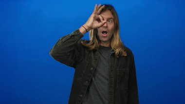 Sarışın, uzun saçlı bir adam mavi stüdyo duvarına ve büzüşen dudaklara karşı tamam işareti tutuyor; merak keşfi oyunculuğu.