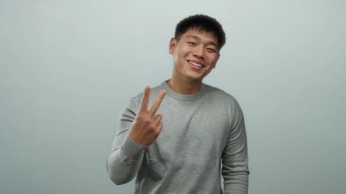 Gri süveter giyen genç Çinli adam izole edilmiş beyaz arka planda elleriyle jestler yapıyor, pozitiflikle gülümsüyor..
