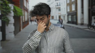 Sakallı genç adam bir sokakta burnunu tutuyor, kötü bir kokuya tepki veriyor, şehir ortamında bir anlık hoşnutsuzluk gösteriyor..