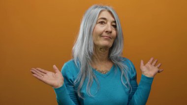 Gri saçlı, mavi kazaklı yaşlı kadın turuncu arka planda hareketler yapıyor.