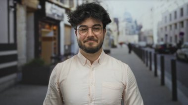 Sakallı genç adam canlı bir şehir caddesinde duruyor, gözlük takıyor ve açık tişört giyiyor, rahat ve şehir dışı yaşam tarzını yansıtıyor..