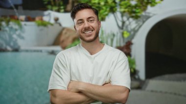 Sakallı genç adam arka planda yemyeşil ve modern mimarisi olan bir otelin havuzunun yanında gülümsüyor..